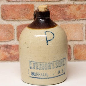 May include: Antique stoneware jug with a cork stopper. The jug is beige with a dark brown top and handle. It features blue lettering that reads "E. PERSON'S SONS, BUFFALO, N.Y." and a blue "P" on the front. The jug is a vintage piece.