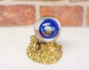May include: A clear glass orb with a swirling blue design, resting on a gold-colored decorative base. The base features a cherub figure and floral details. The background includes a brick wall.