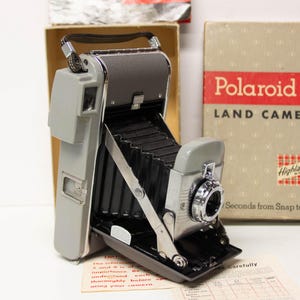 May include: A vintage Polaroid Land Camera Highlander Model 80A with a gray body and black bellows. The camera is positioned in front of its original cardboard box, which features the text Polaroid LAND CAMERA and Highlander Model 80A.