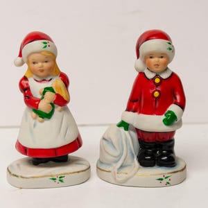 May include: Two vintage porcelain Christmas figurines. A girl in a red dress and Santa hat holds a doll. A boy in a red suit and Santa hat carries a sack. Both figurines stand on a white base with green holly.
