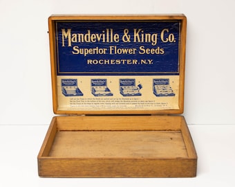May include: A rectangular wooden box with an open lid featuring a blue and cream label that reads Mandeville & King Co. Superior Flower Seeds Rochester, N.Y. The interior lid displays four instructional diagrams and text for seed display setup.