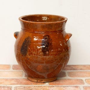 May include: A round, glazed earthenware pot with a wide mouth and two small side handles. The vessel features a glossy, deep amber-brown finish with darker, mottled brown spots distributed across the exterior surface and a visible crackle texture.
