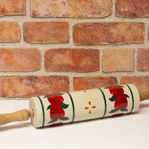 May include: A white rolling pin with a wooden handle. The rolling pin is decorated with red apples and green leaves on a white background.