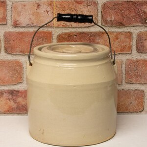 May include: A beige stoneware crock with a lid and a black wooden handle. The crock has a metal wire handle and is set against a brick wall background. The antique kitchenware is a vintage piece.