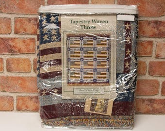 May include: Tapestry woven throw featuring an American flag design with red, white, and blue colors. The throw is packaged in plastic and has a label that reads "Tapestry Woven Throw" and "Americana Flag".