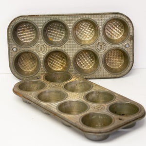 May include: Two vintage metal muffin tins. The top tin has six round muffin cups and the bottom tin has eight round muffin cups. Both tins are silver with a textured surface.
