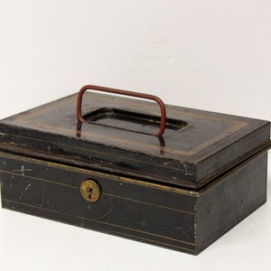 May include: A rectangular black metal cash box featuring a thin red wire handle on the lid. The box has a brass-colored keyhole on the front panel and is decorated with subtle, thin gold-toned pinstripe lines along the edges of the lid and body.