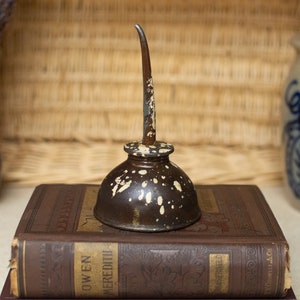 May include: A vintage oil can with a long spout, painted in brown and white with a speckled pattern. The can is sitting on top of two antique books.