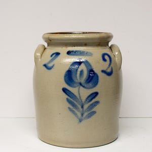 May include: A tan stoneware crock with two small handles and a blue floral design. The crock features a blue flower with leaves and the number "2" on either side of the flower. The crock is a vintage piece.