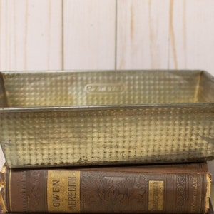 May include: Vintage rectangular metal loaf pan with a textured surface, resting on top of two antique books. The pan has a slightly aged appearance, suggesting it has been used for baking. The books have gold lettering.