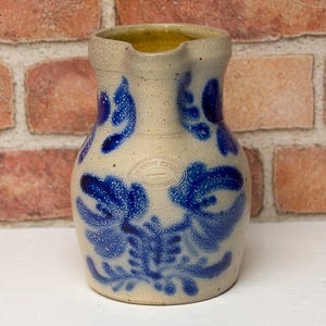 May include: A stoneware pitcher with a light beige base and vibrant blue floral designs. The pitcher has a spout and a handle, and the interior is a warm yellow. The design is reminiscent of antique pottery.