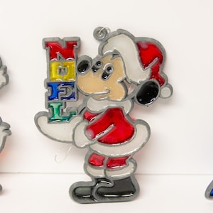 May include: Set of three Christmas ornaments featuring cartoon characters. Mickey Mouse in a Santa suit holding a "NOEL" sign, Donald Duck, and a character in blue and white attire. Each ornament has a silver outline and colorful accents.