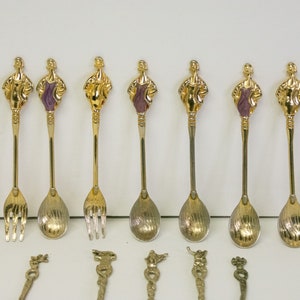 May include: Set of eight gold-tone teaspoons with a purple and gold design. The spoons feature a woman figure design. The set also includes four additional gold-tone teaspoons with different designs.
