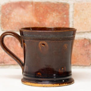 May include: A dark brown ceramic mug with a curved handle. The mug has a cylindrical shape with a decorative band near the top and a lighter-colored base. The background features a brick wall.