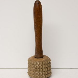 May include: A wooden meat tenderizer with a dark brown handle and a light brown, textured head. The head features a grid of raised, pyramid-shaped bumps. The handle is smooth and cylindrical, with a hole at the top.