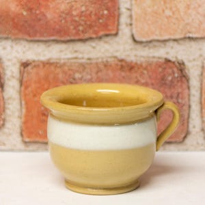 May include: A small, ceramic mug with a handle, featuring a two-tone design. The top and bottom sections are a warm yellow, while a white band encircles the middle. The mug is set against a brick wall background.