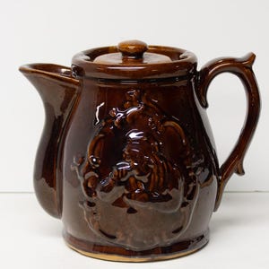 May include: A brown ceramic teapot with a lid and handle. The teapot has a glossy finish and a relief design on the front. The spout is curved, and the handle is arched. A classic kitchenware item.
