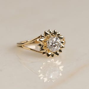Peut inclure: Une bague en or avec une pierre précieuse ronde et transparente au centre, entourée d'un halo de pétales en or disposés en forme de fleur. L'anneau est divisé en deux segments fins en or poli qui se rejoignent à la base du sertissage.