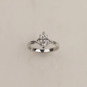 Peut inclure: Une bague argentée ornée d'une pierre transparente unique taillée en marquise et fixée par des griffes. L'anneau présente un design divisé qui converge à la base de la pierre, offrant une apparence lisse et polie avec une finition métallique.