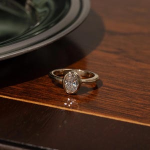 May include: A gold ring featuring a single oval-cut clear gemstone set in a smooth bezel setting. The band is polished and rounded, showcasing a clean and simple design that highlights the central stone's clarity and the warm metallic luster.