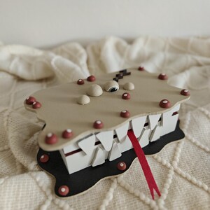 May include: A rectangular box with a tan lid featuring small red and white dots, a single eye, and a black lightning bolt. The box has white triangular teeth along the sides, a red tongue protruding from the front, and a black base with red dots.
