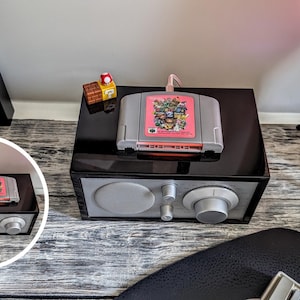 Può includere: Una docking station per altoparlanti rettangolare nera con pannello frontale argentato, una grande manopola circolare per il volume e due manopole di controllo più piccole. Una cartuccia per videogiochi grigia con etichetta rosa è inserita.