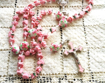 May include: A pink and white rosary with a silver crucifix. The beads are shaped like roses and have green leaves. The crucifix has a pink enamel finish.