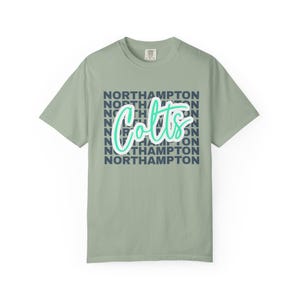 May include: A sage green short-sleeve t-shirt featuring the word Colts in a bright teal script font layered over the word NORTHAMPTON repeated in dark navy blue block letters across the center of the chest. The shirt has a crew neckline.