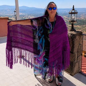 Pode incluir: Um xale de crochê roxo vibrante com um padrão texturizado de malha aberta e franjas longas e fluidas na borda inferior. O xale está drapeado sobre um vestido preto semitransparente com um padrão abstrato colorido em tons de azul e rosa.