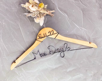 Könnte beinhalten: Ein Holz Kleiderbügel mit einer Drahtinschrift, die "Mrs. Doyle 6.11.22" lautet. Der Kleiderbügel ist mit getrockneten Blumen und Grünpflanzen verziert.