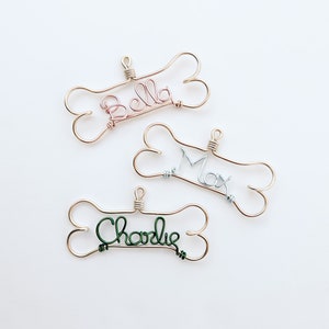 May include: Three handmade dog bone ornaments with names. Each bone is crafted from gold-colored wire, with the names spelled out in pink, silver, and green wire. The names are "Bella", "Max", and "Charlie".