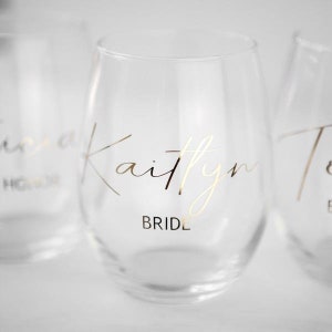 Puede incluir: Tres copas de vino de vidrio transparente con letras doradas. La copa del centro dice "Kaitlyn Bride".