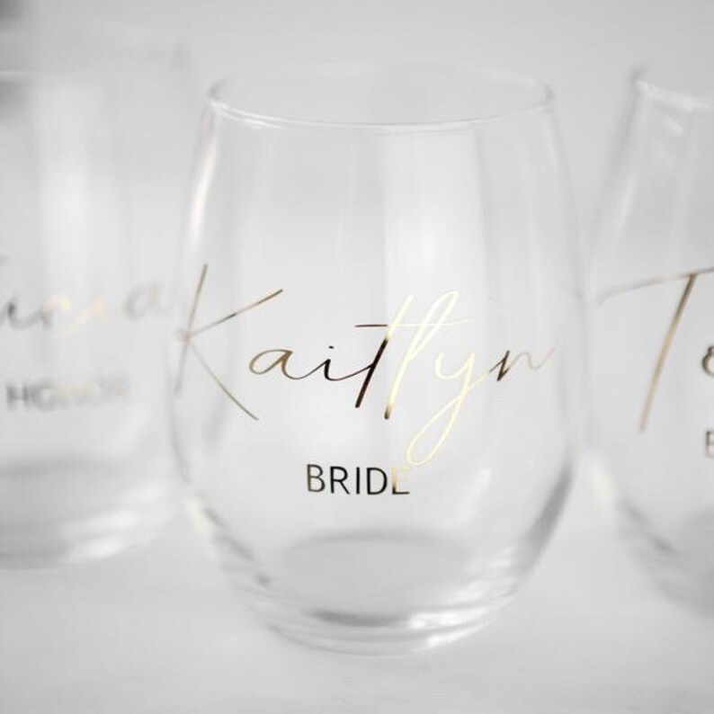 Puede incluir: Tres copas de vino de vidrio transparente con letras doradas. La copa del centro dice "Kaitlyn Bride".