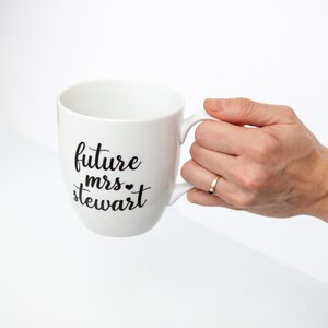 以下が含まれることがあります： 白いセラミック製のマグカップに、黒い文字で「future mrs. stewart」と小さなハートマークが書かれています。