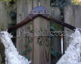 May include: A brown wooden hanger with a silver wire inscription that reads "Mrs. Gutierrez" and "10.5.13". A white lace dress is hanging on the hanger.