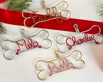 May include: Four wire dog bone ornaments with the names Buddy, Milo, Charlie, and Bella written in red, pink, and copper wire.