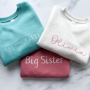 May include: Three sweatshirts stacked on top of each other. The top sweatshirt is white with the name "Olivia" embroidered in pink. The middle sweatshirt is teal with the word "custom" embroidered in white. The bottom sweatshirt is pink with the words "Big Sister" embroidered in white.