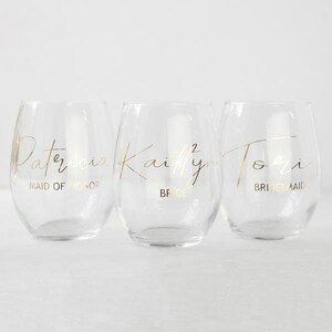 Puede incluir: Tres copas de vino de vidrio transparente con letras doradas. Las copas est&aacute;n etiquetadas como "Patricia Maid of Honor", "Kaitlyn Bride" y "Tori Bridesmaid".