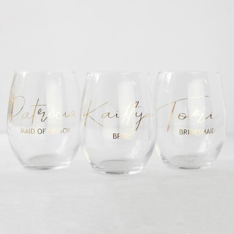 Puede incluir: Tres copas de vino de vidrio transparente con letras doradas. Las copas est&aacute;n etiquetadas como "Patricia Maid of Honor", "Kaitlyn Bride" y "Tori Bridesmaid".