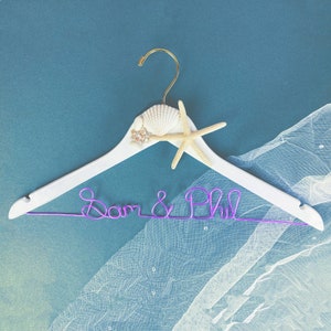 May include: White wooden hanger with a seashell and starfish design. The hanger has a purple wire inscription that reads "Sam & Phil".