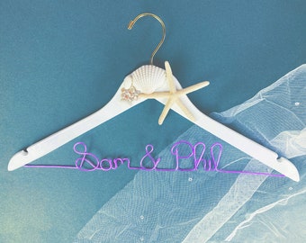 May include: White wooden hanger with a seashell and starfish design. The hanger has a purple wire inscription that reads "Sam & Phil".