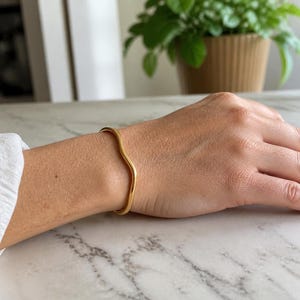 May include: A gold-toned metal bangle bracelet featuring a smooth, continuous wavy or undulating design. The bracelet has a polished, reflective surface and is worn around the wrist, showcasing a simple, curved silhouette without any additional embellishments.