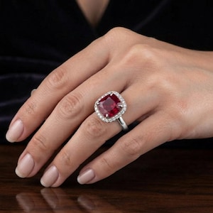 Könnte beinhalten: Ein silberner Ring mit einem großen, kissenförmigen, tiefroten Rubin in der Mitte, umgeben von einem Kranz aus kleinen, klaren, rund geschliffenen weißen Diamanten auf einer polierten Silberschiene, die zum Finger hin schmaler wird.
