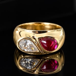 May include: A wide gold band ring featuring two pear-shaped gemstones set side-by-side. One gemstone is a clear, faceted white stone, and the other is a deep red, smooth cabochon-style stone. The gold band has a polished, reflective finish.