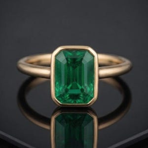 Peut inclure: Une bague en or avec une pierre précieuse verte taille émeraude sertie dans un chaton lisse. L'anneau est en or poli avec un profil arrondi, mettant en valeur une pierre verte vive qui se reflète nettement sur la surface sombre en dessous.