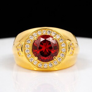 Puede incluir: Un anillo de oro con una gran piedra preciosa de color rojo intenso, redonda y facetada en el centro. La piedra central está rodeada por un halo de pequeñas piedras blancas claras y brillantes engastadas en la banda de oro, que también incluye piedras laterales.
