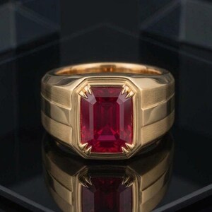 May include: A gold ring featuring a large, rectangular, emerald-cut red gemstone set in the centre. The band has a wide, brushed metal finish with horizontal grooved detailing on the sides, creating a bold and structured appearance.