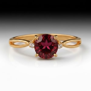 Könnte beinhalten: Ein Goldring mit einem runden, tiefroten Edelstein in der Mitte, flankiert von zwei kleinen, klaren Akzentsteinen. Die Ringschiene hat ein geteiltes Bypass-Design, das sich zum Stein hin verjüngt, gefertigt aus poliertem Gelbgold.