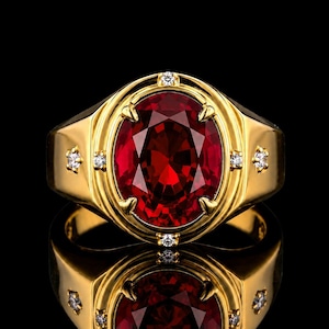Peut inclure: Une bague en or ornée d'une grande pierre précieuse rouge foncé de taille ovale au centre. L'anneau est agrémenté de quatre petites pierres claires de taille ronde incrustées dans le métal doré de chaque côté du sertissage central.