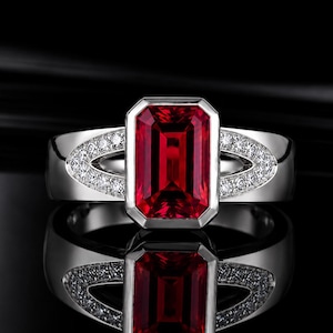 May include: A silver-toned ring featuring a large, rectangular, emerald-cut red gemstone at the centre. The band is accented with small, clear, round-cut stones set in a V-shaped pattern on both sides of the central red stone setting.
